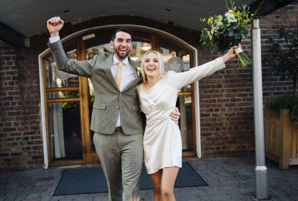 Celebratory exit pose under brick archway with couple in modern wedding attire.