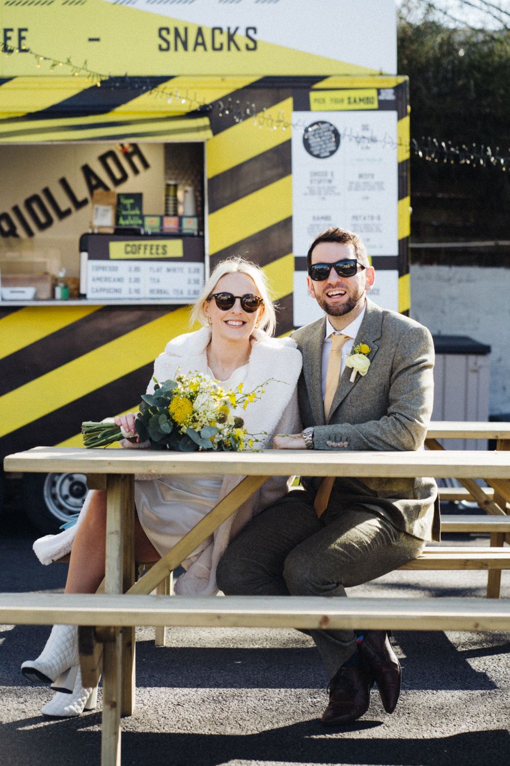 A couple in sunglasses sitting at a yellow food truck with wedding flowers and casual style.