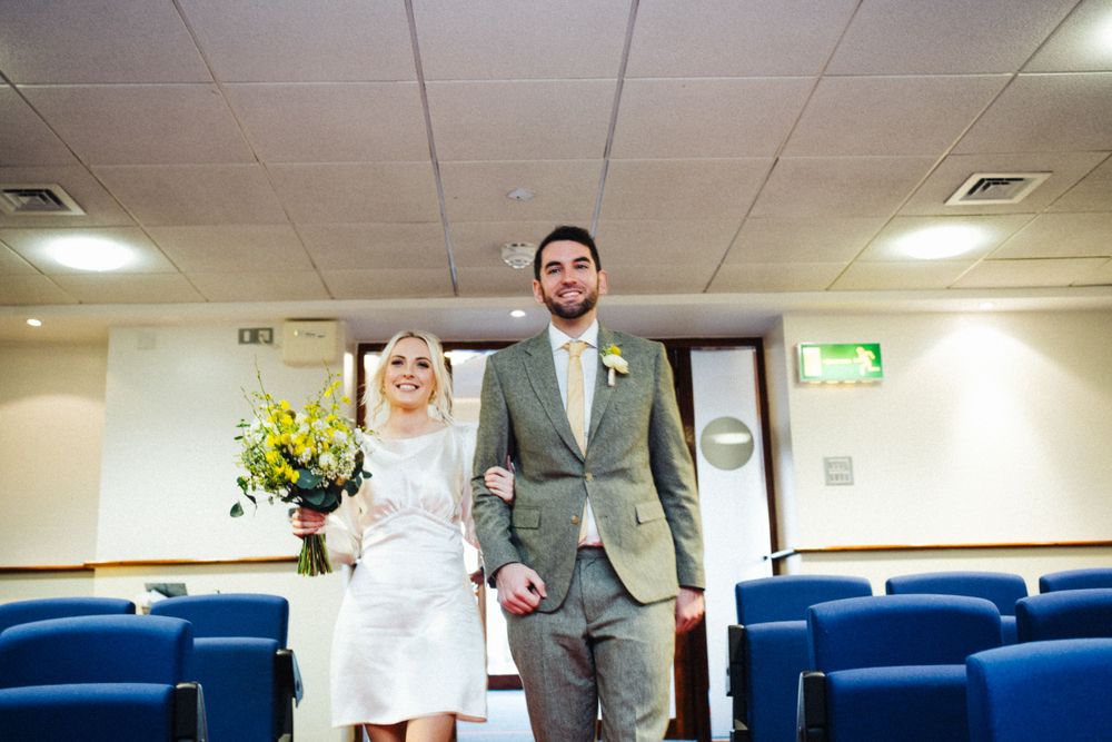 A wedding couple standing together in a simple room with blue chairs and fluorescent lighting.