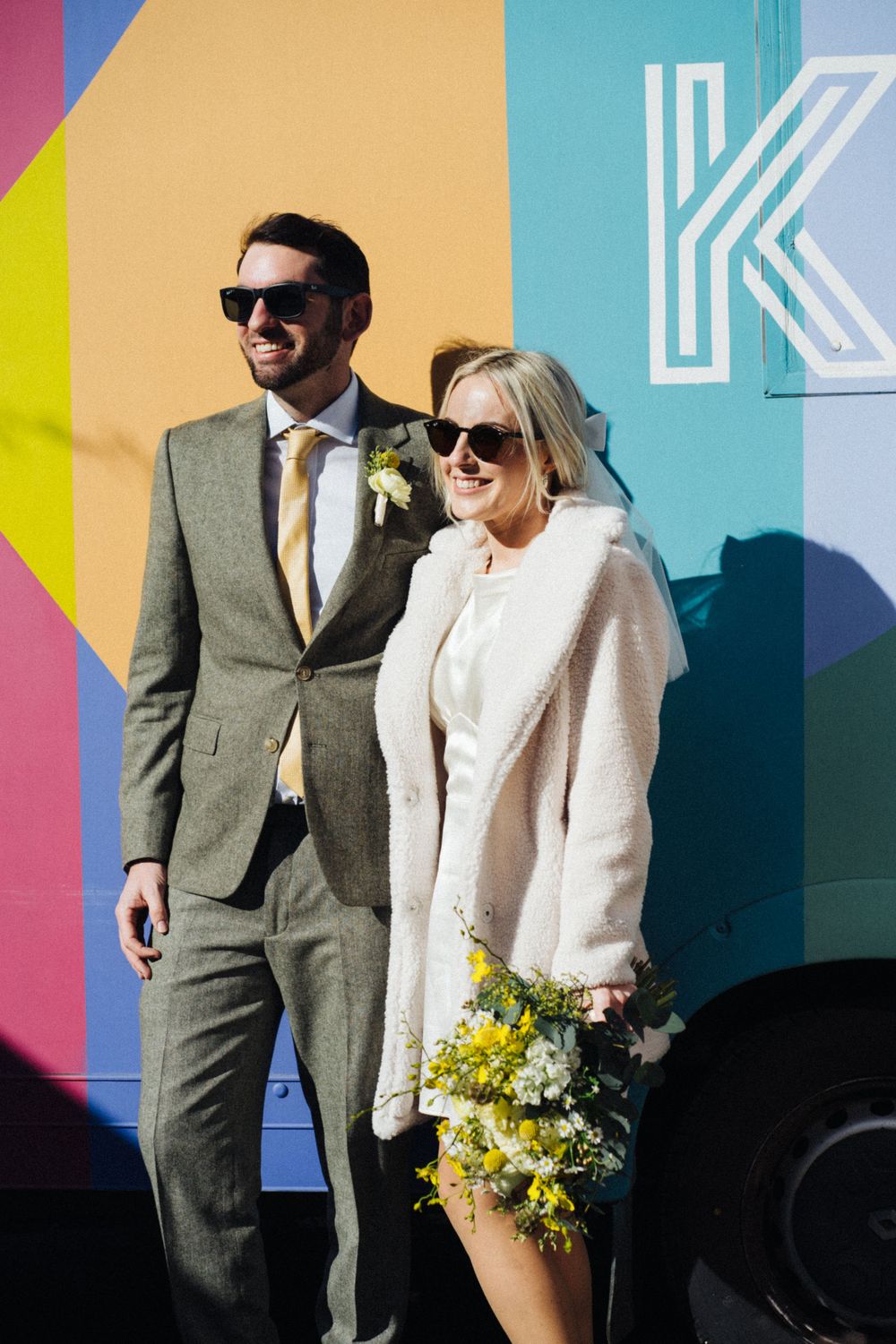 Stylish couple poses against colorful geometric mural wall with yellow flowers and white boots.