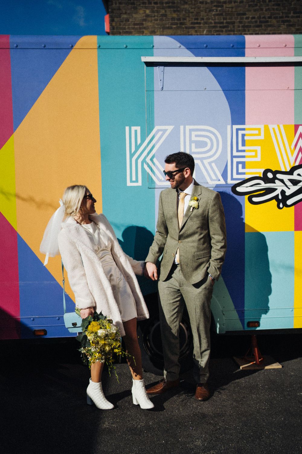 Modern wedding couple standing by vibrant street art with yellow bouquet and white boots.