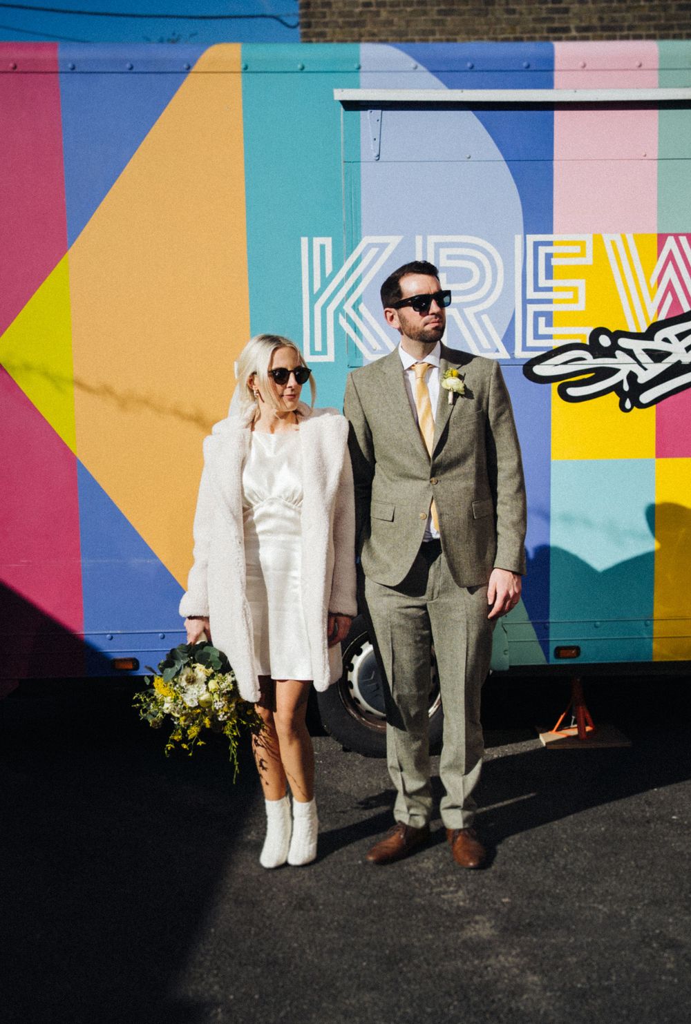 Fashionable couple in sunglasses poses by colorful graffiti wall with yellow floral bouquet.