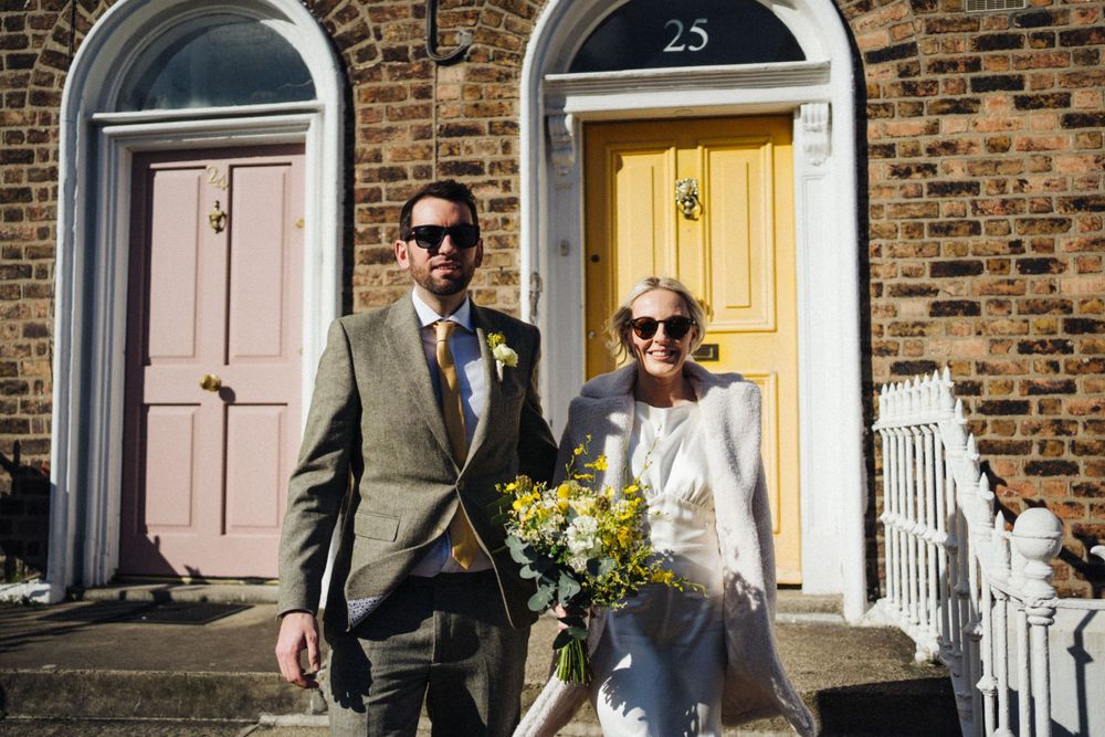 Couple in formal attire poses on steps of brick townhouse with yellow door.
