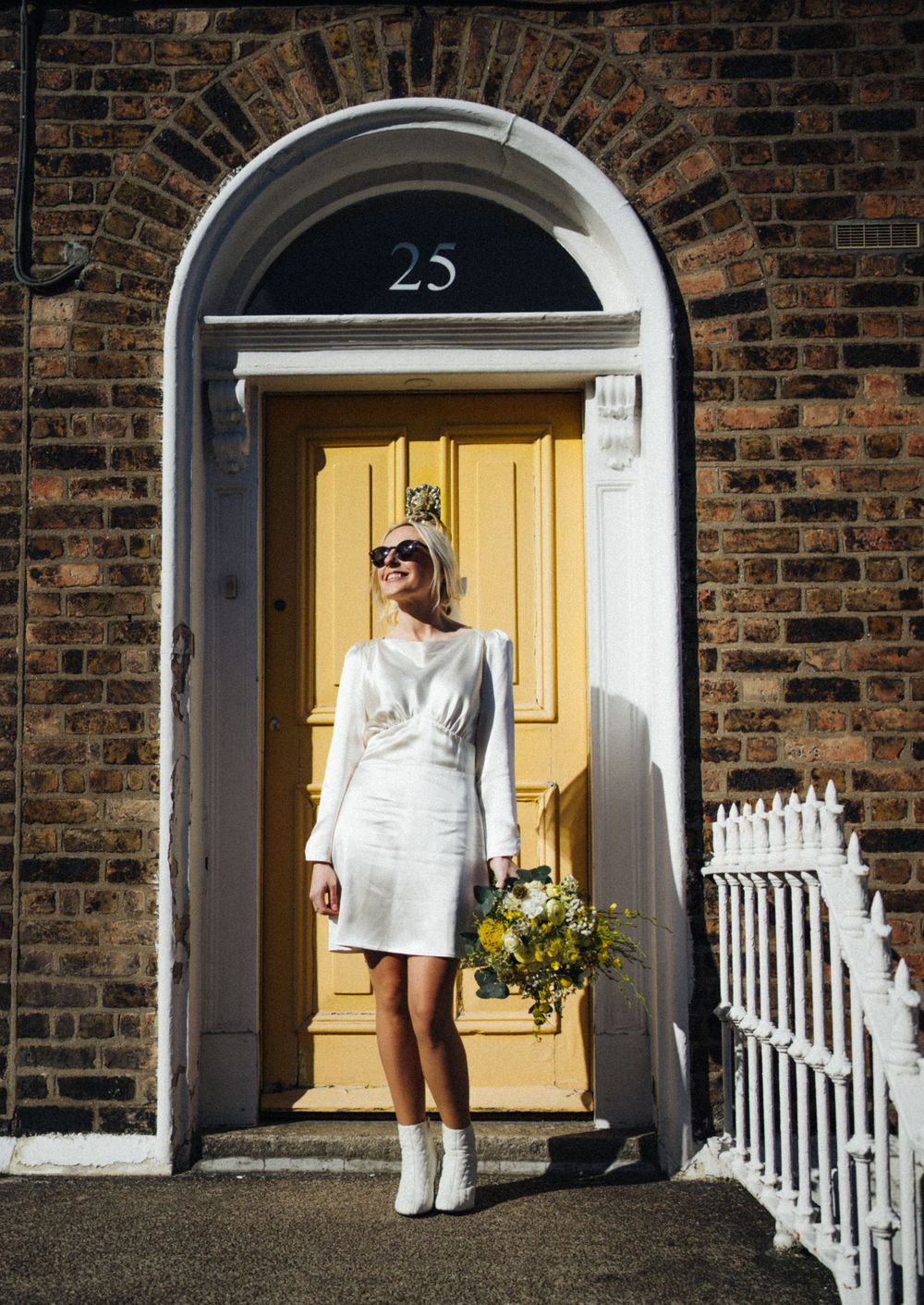 Fashionable figure in white dress and boots stands in yellow doorway of brick building.