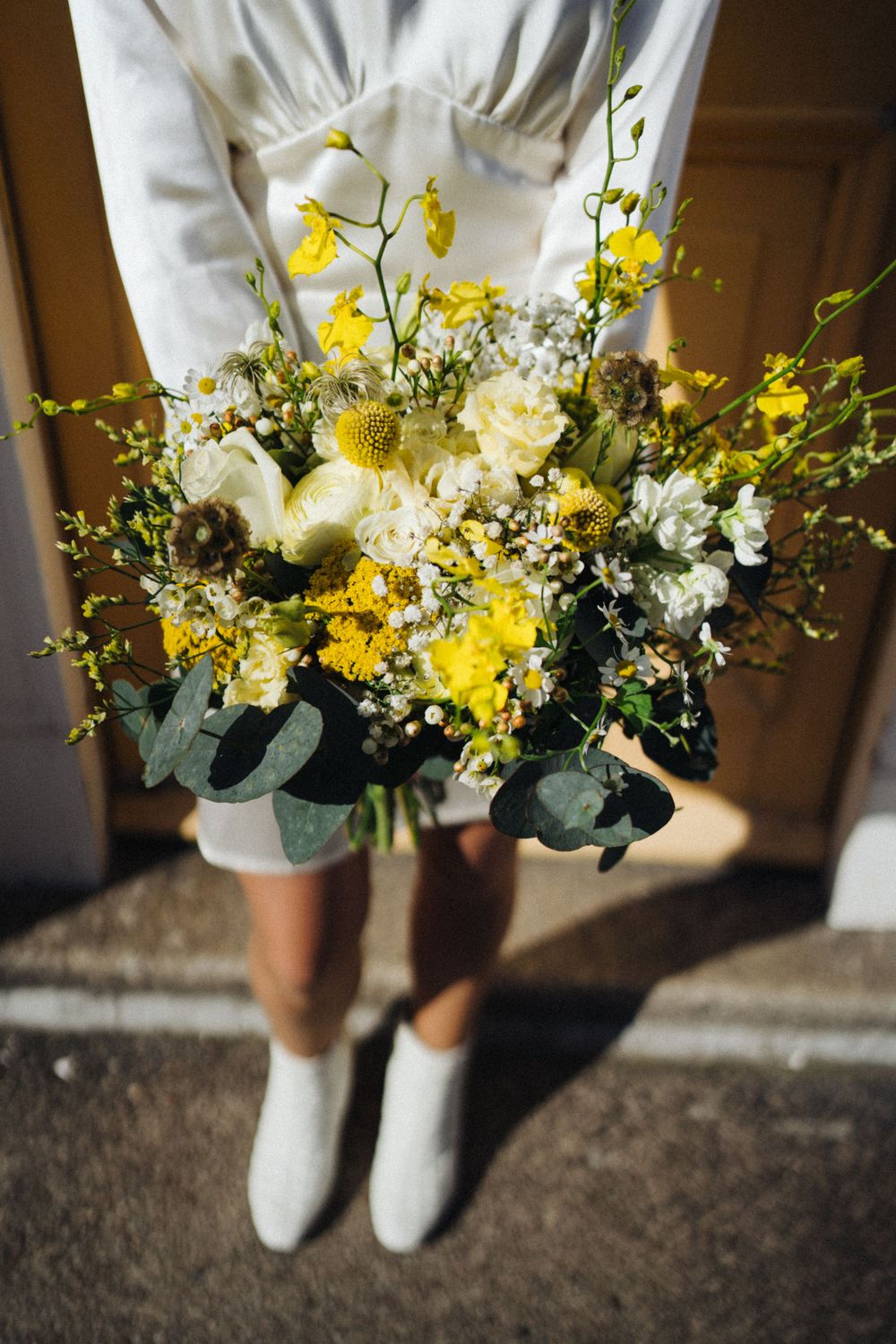 Yellow and white floral bouquet with eucalyptus held against white dress and boots.
