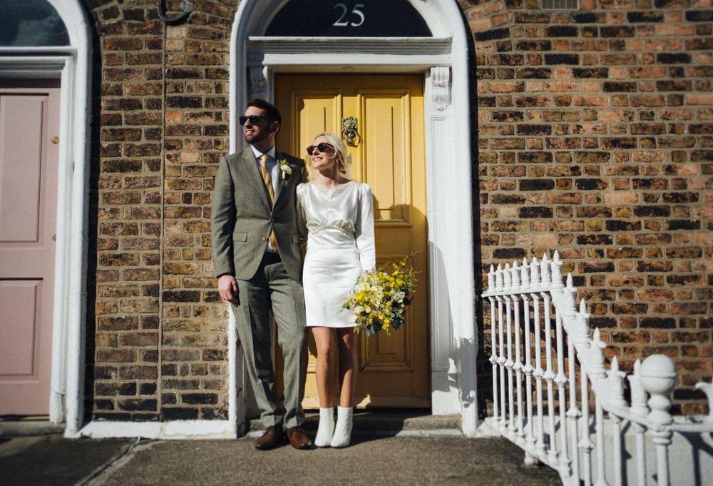 Elegant couple poses in doorway of brick building with yellow flowers and white boots.