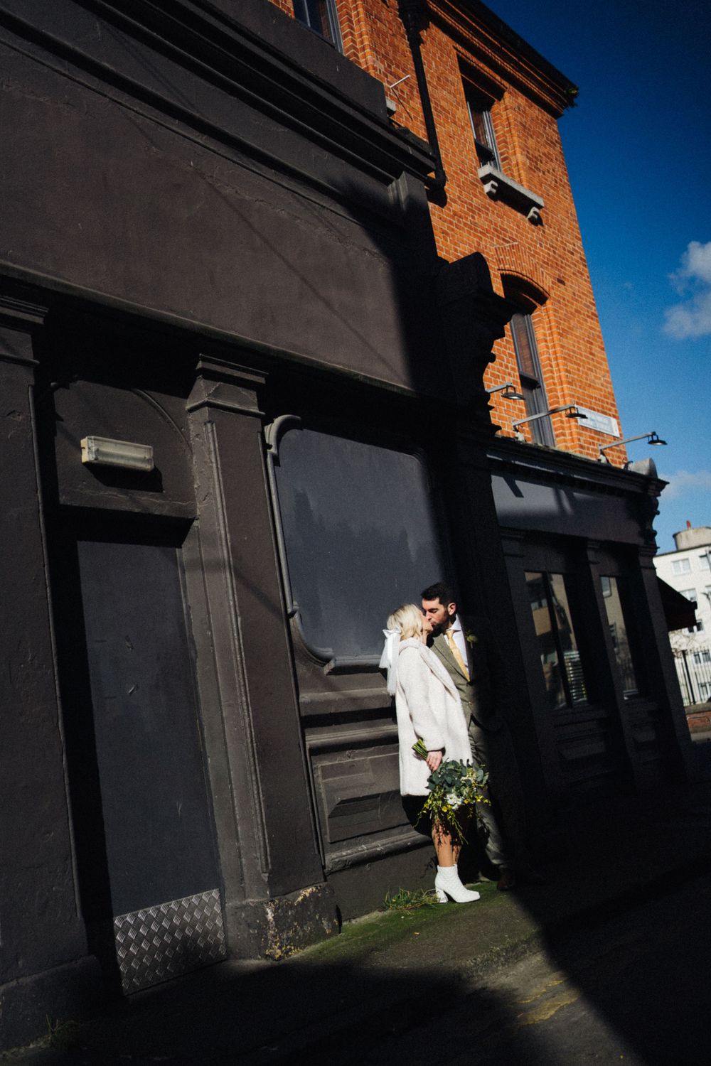 Couple in formal wear stands against black storefront of brick building.