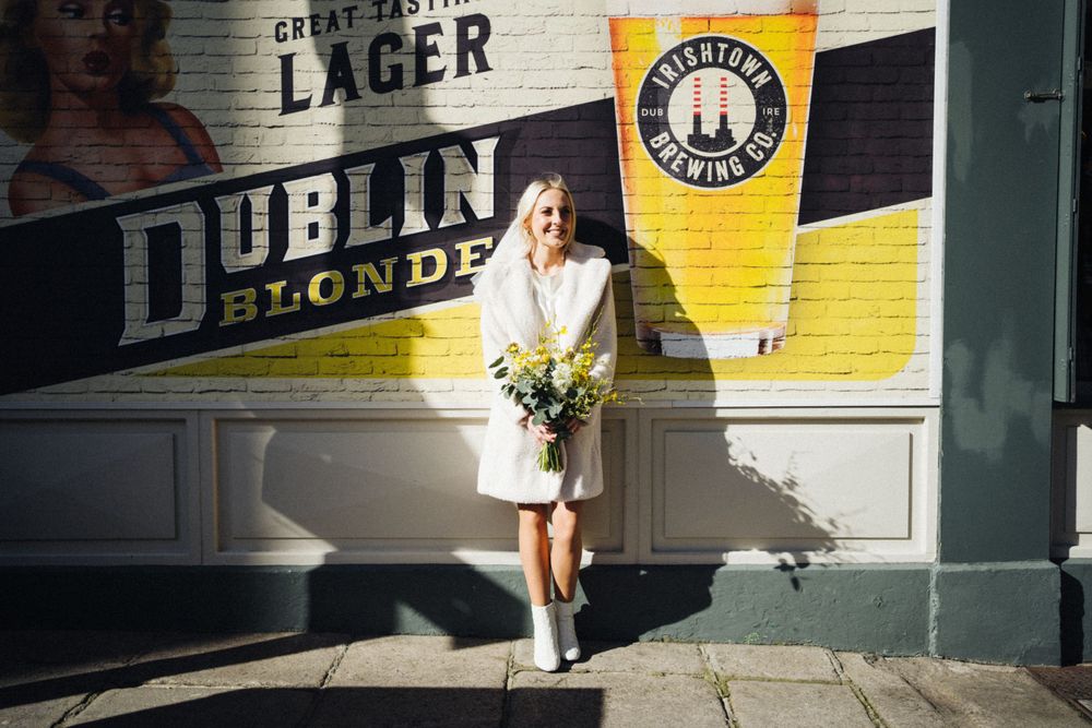 Figure in white dress holds yellow bouquet against Dublin Lager advertisement wall.