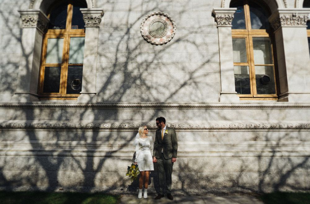 A couple stands against a white wall with ornate windows casting dramatic tree shadows.