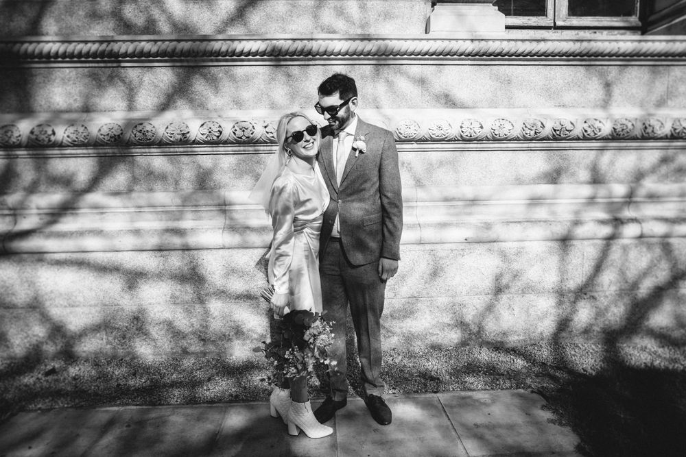 A couple poses against a white wall with dramatic shadow patterns in black and white photography.