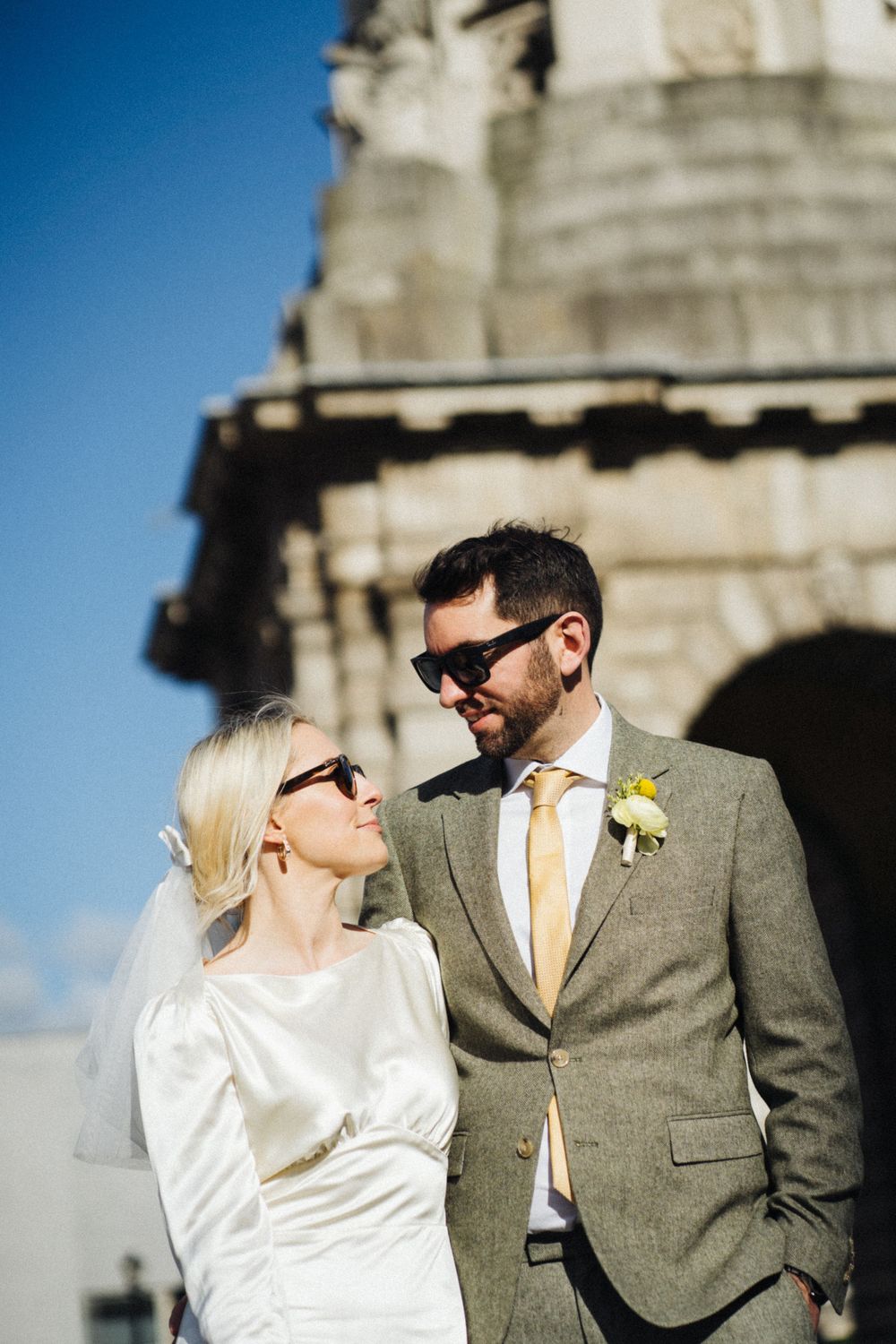 A couple in stylish wedding attire poses in front of historic architecture on a sunny day.