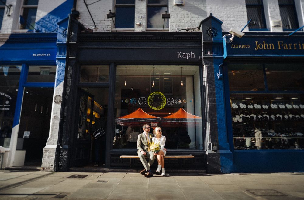A couple sits outside a blue-trimmed storefront with neon signage.