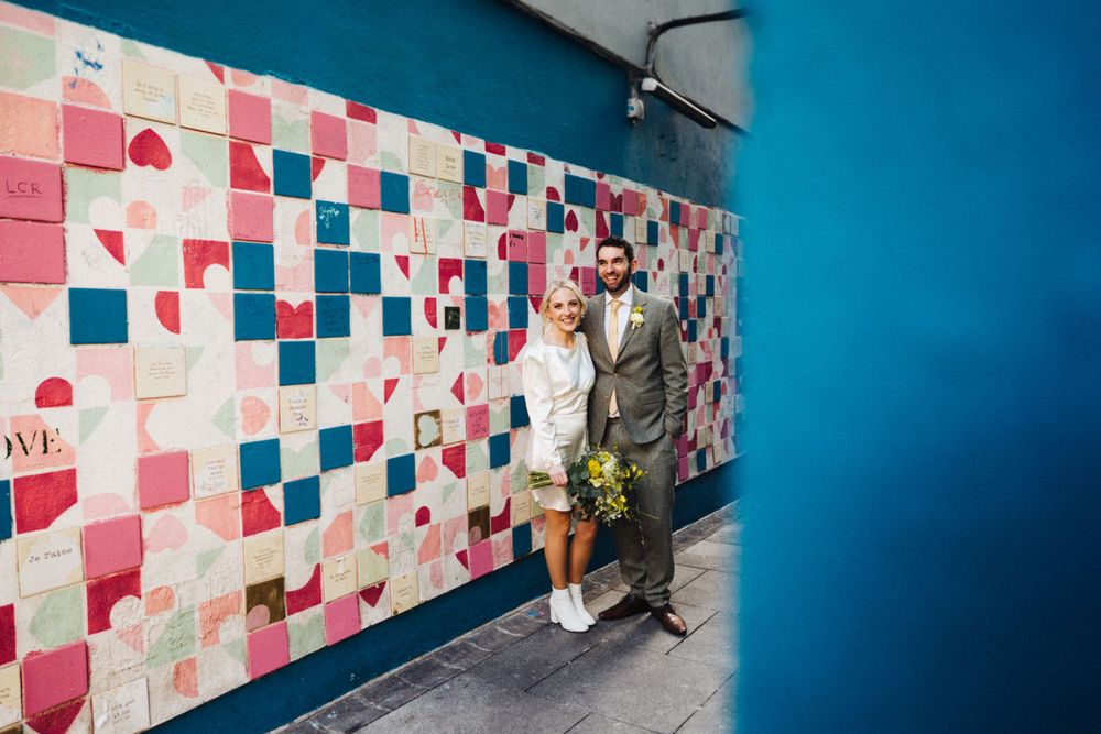 A couple poses against a colorful patchwork wall design in an urban setting.