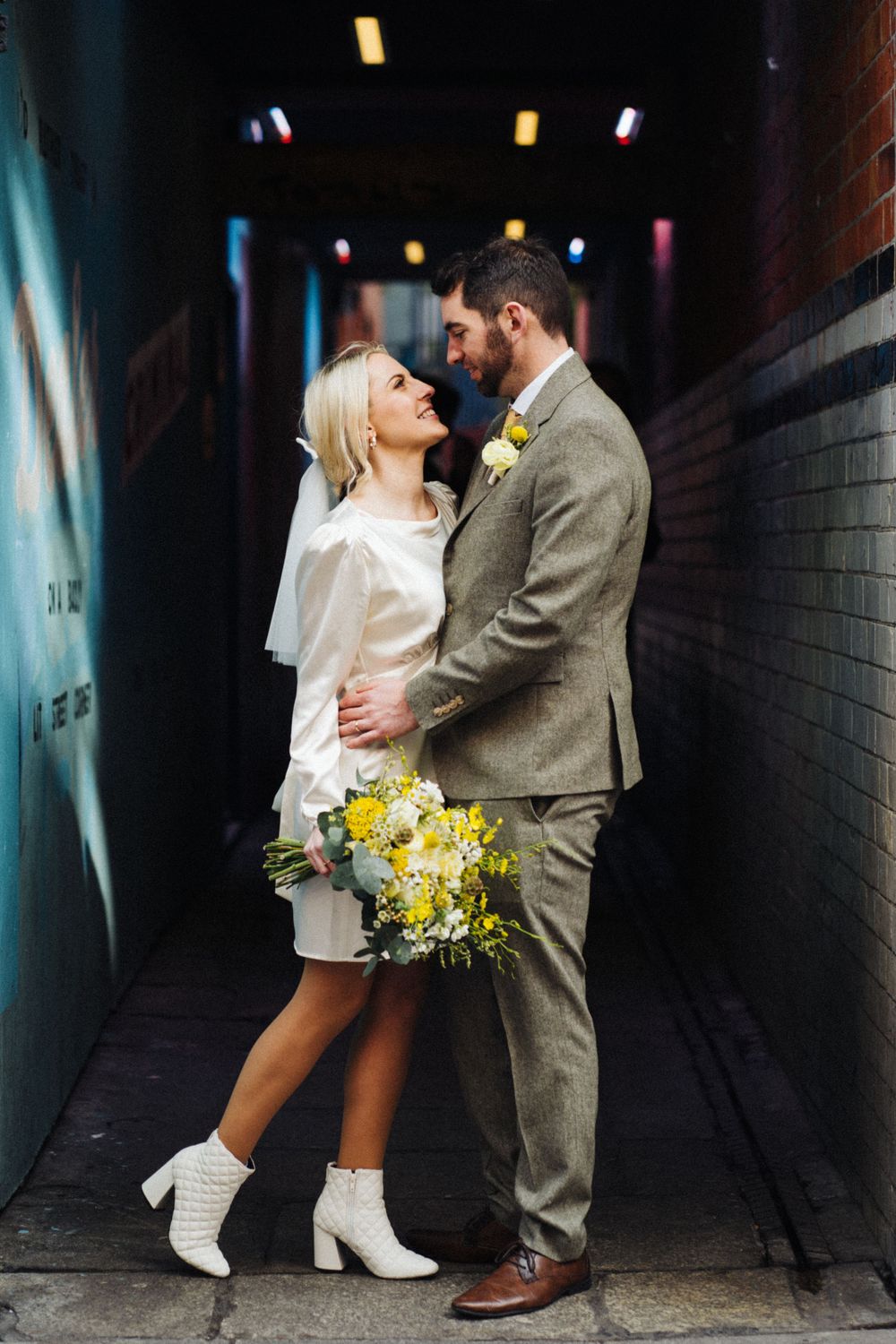 A tender moment between a couple in white and grey attire holding yellow flowers in an alley.