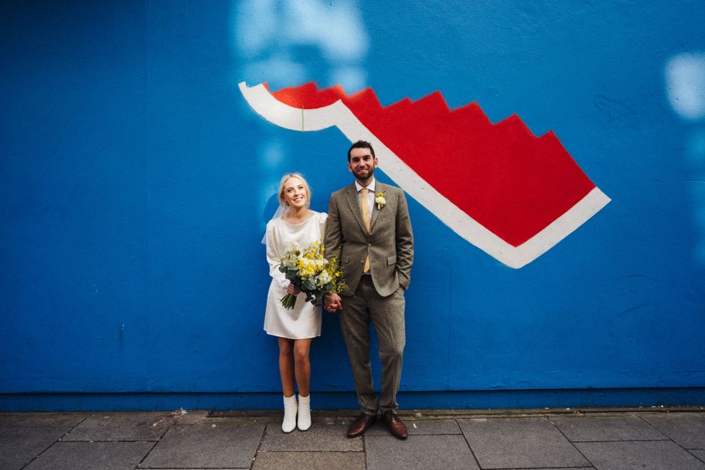 A couple stands against a bright blue wall with a red geometric design.