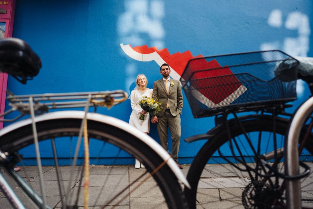 A miniature wedding scene creatively framed through a bicycle wheel against a blue wall.