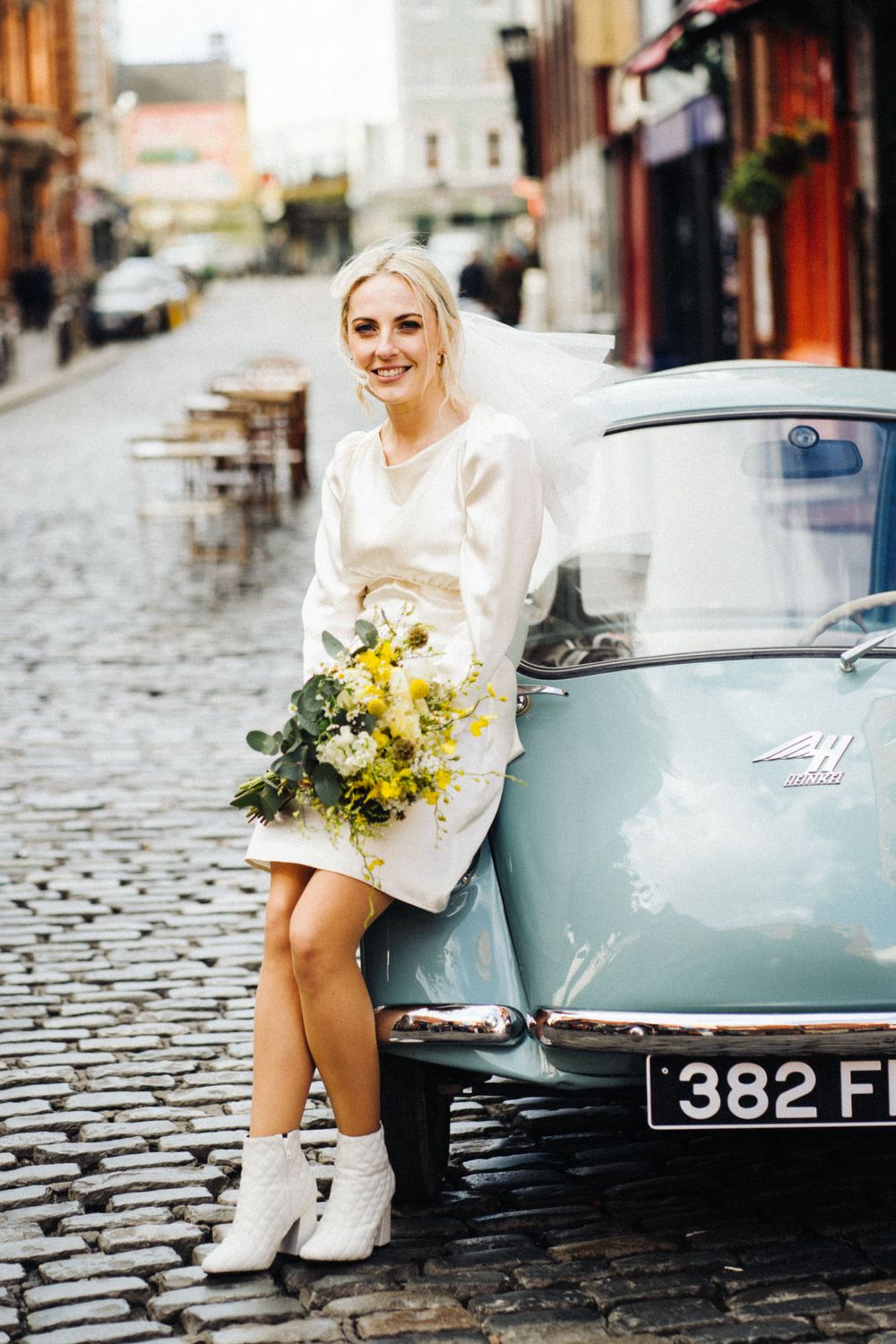 A bride in a short white dress and boots poses with a yellow bouquet next to a vintage blue car on a cobblestone street.