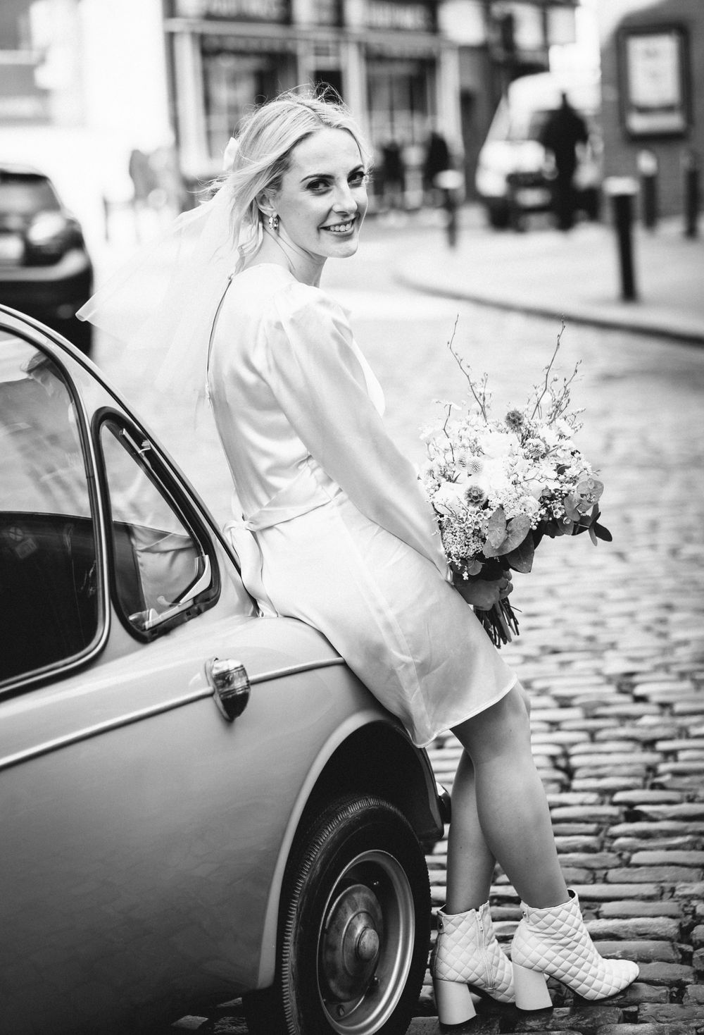 A black and white photo of a bride in a short dress leaning against a vintage car on cobblestone streets.
