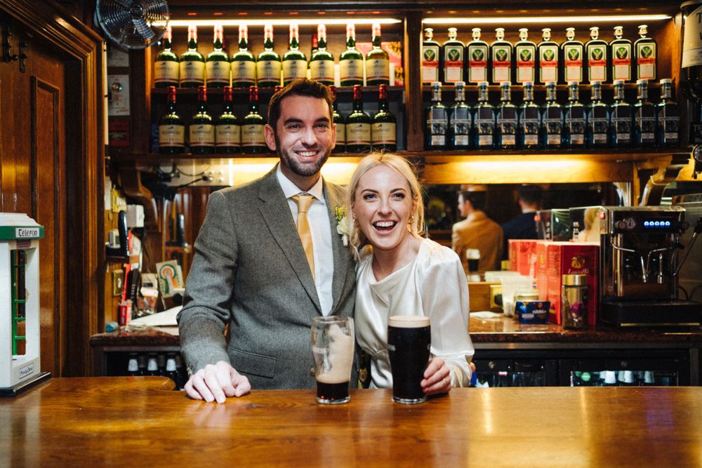 A couple posing behind a bar counter with glasses of dark beer and illuminated bottle display.