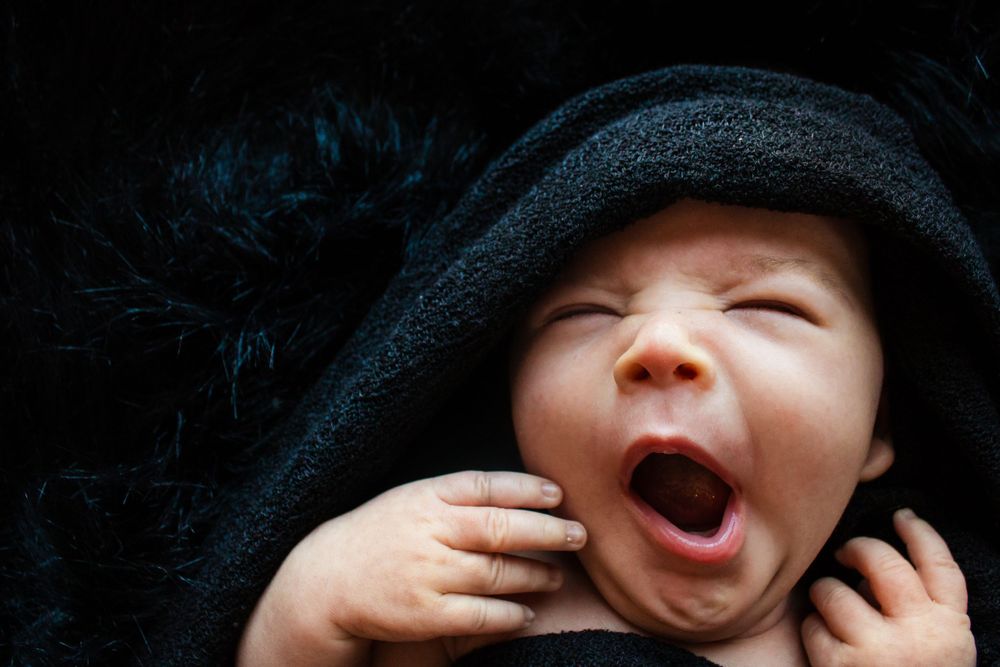 A tiny baby in a black wrap yawns widely while touching their face.