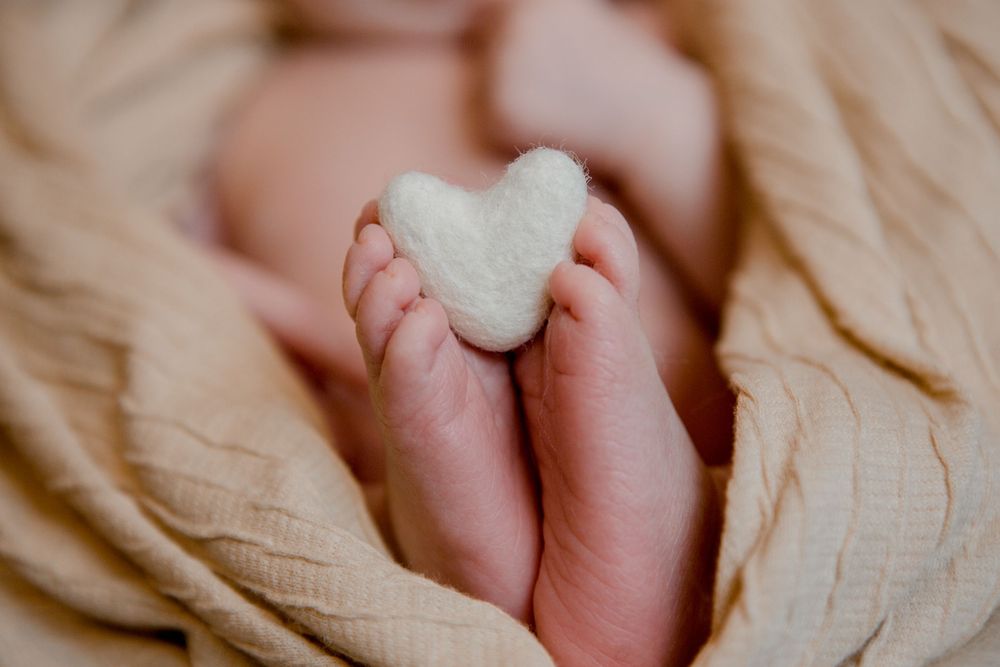 Tiny baby feet holding a small white heart shape against cream fabric.