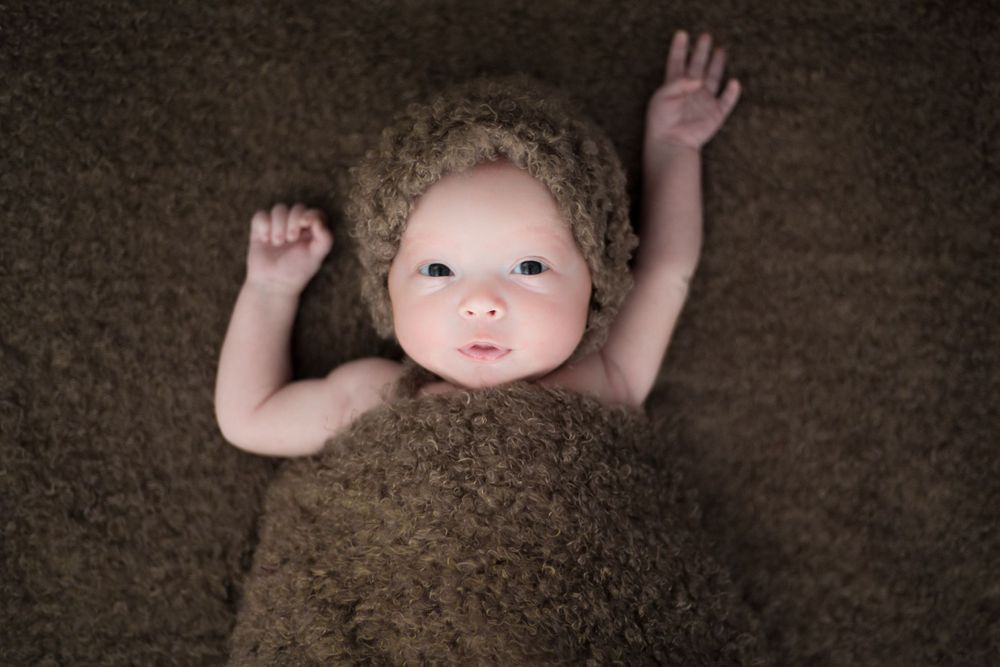 A baby lies on brown fabric with arms raised during a portrait session.