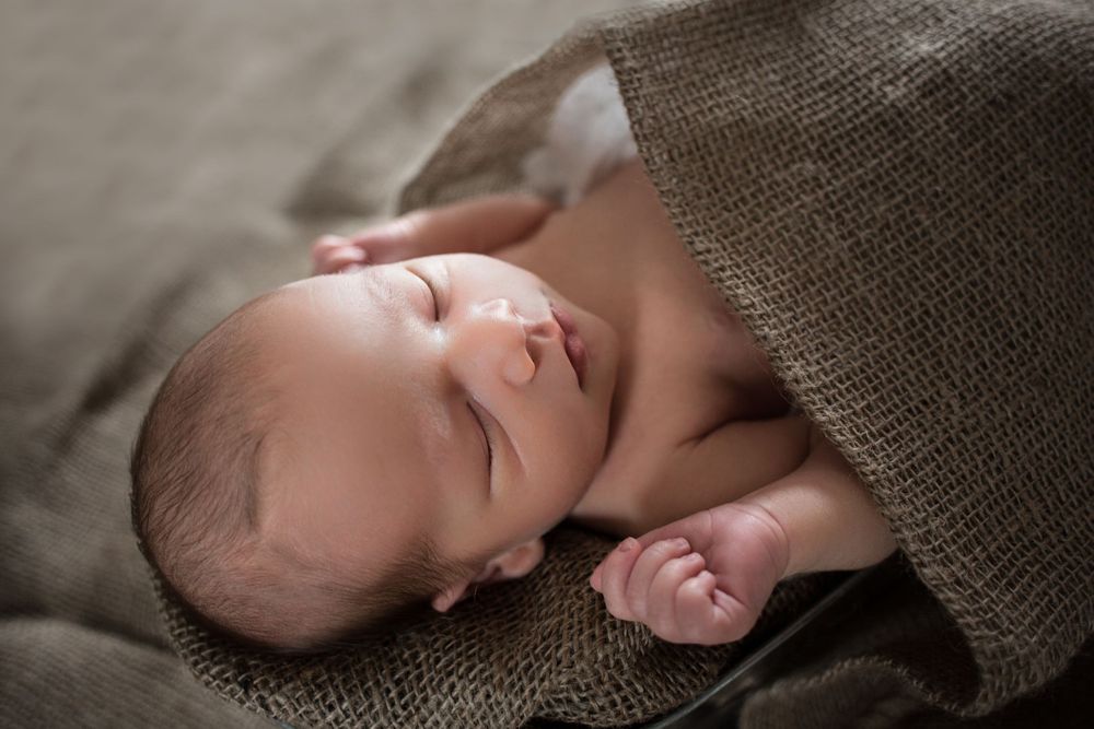 A sleeping infant lies wrapped in a brown blanket during a newborn photography session.