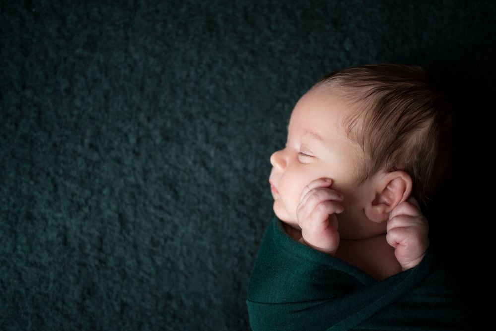 A newborn wrapped in dark green fabric sleeps peacefully against a textured green background.