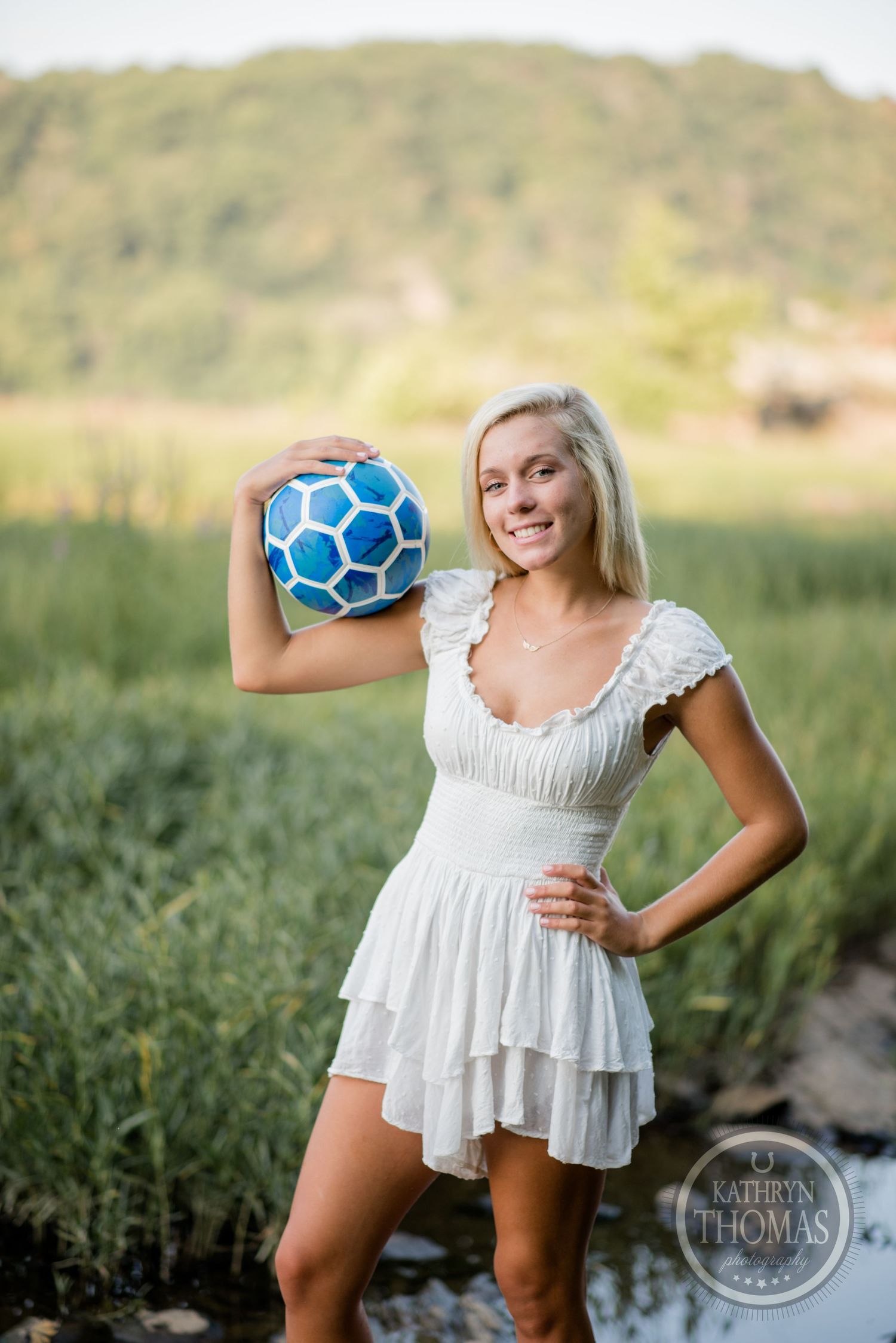 Woman in white sundress holds a blue soccer ball while posing in a grassy field.