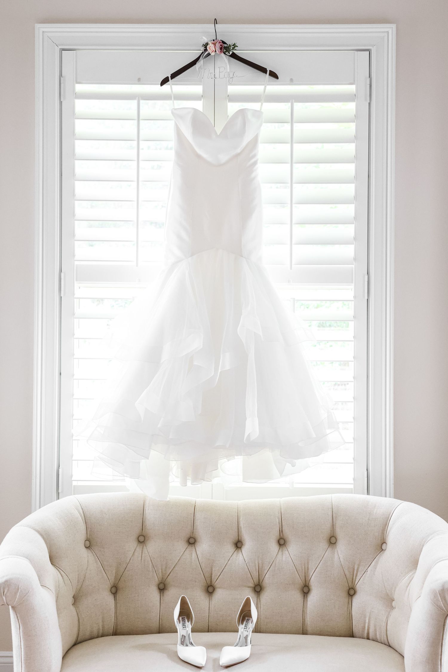 Elegant white wedding dress hanging in window light above tufted cream sofa.