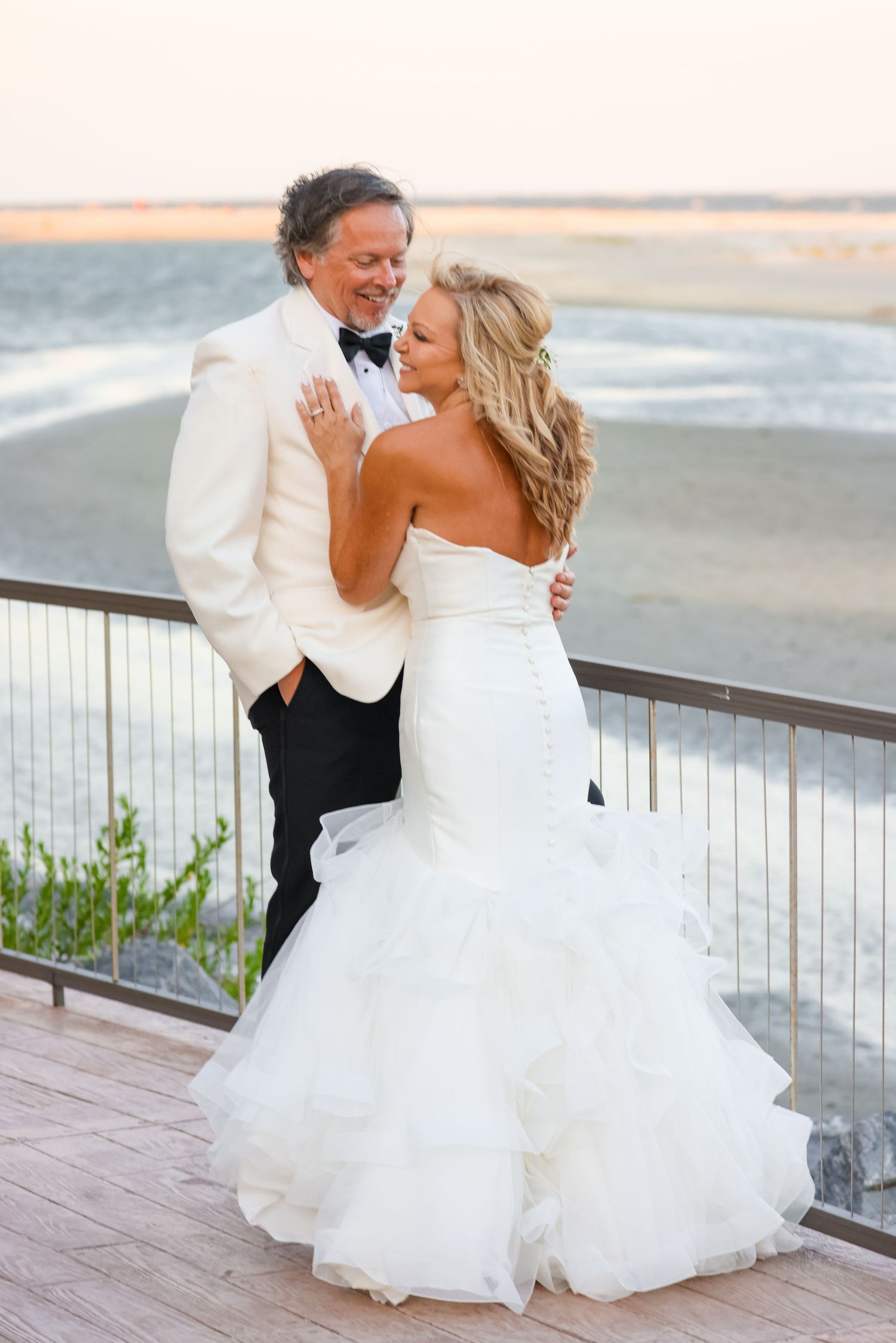 A romantic wedding couple embraces on a beach deck at sunset, with the bride wearing a flowing white mermaid gown.