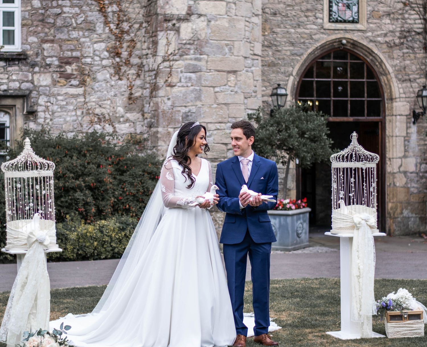 Wedding couple photographed along stone wall with decorative bird cages and floral arrangements.