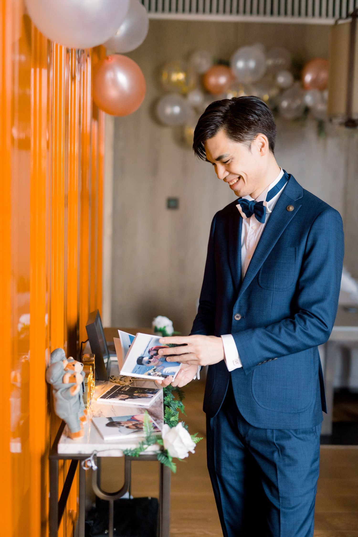 Groom in navy suit reads a letter surrounded by white and peach balloons.