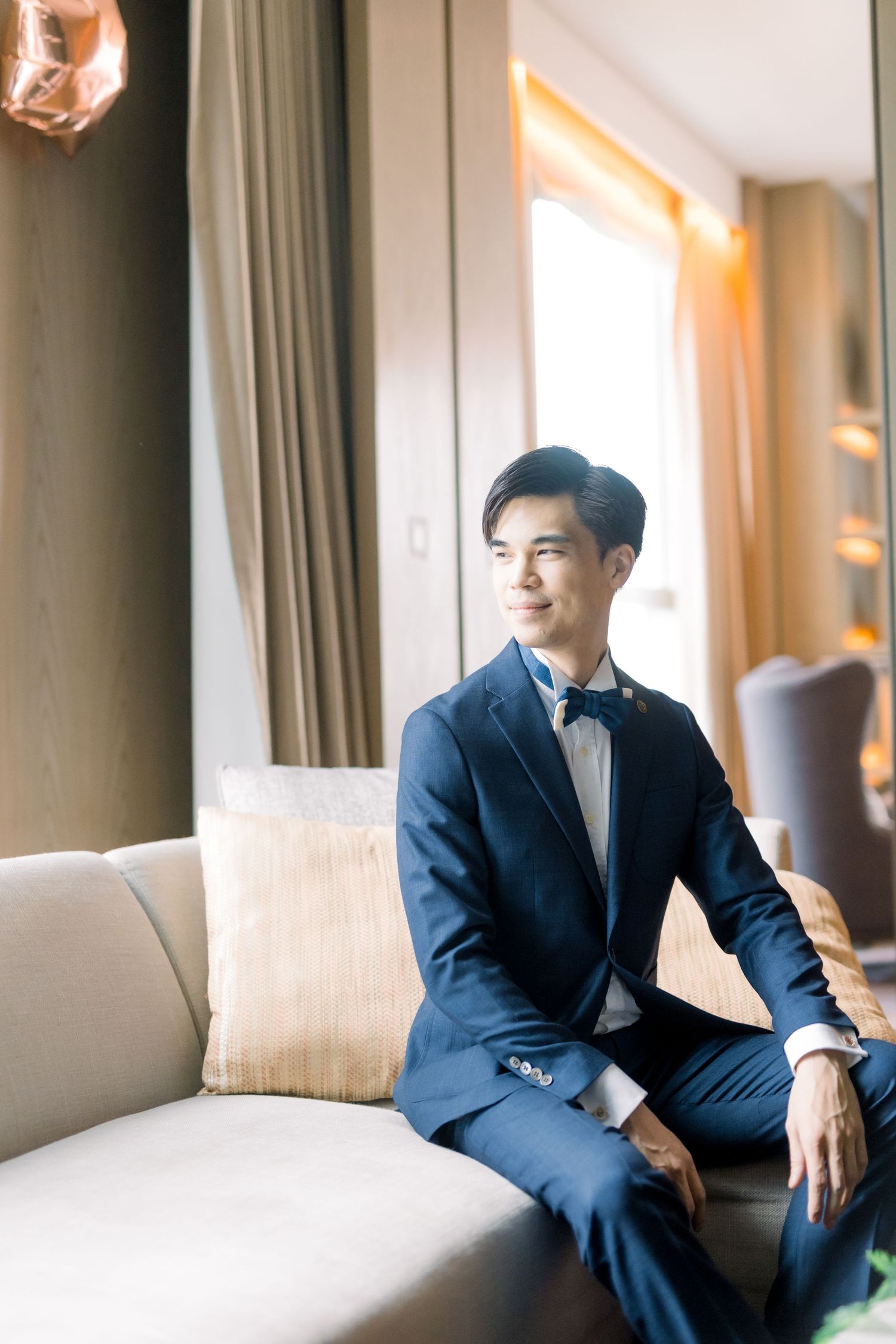 Groom in formal navy suit relaxes on beige couch in sunlit room.