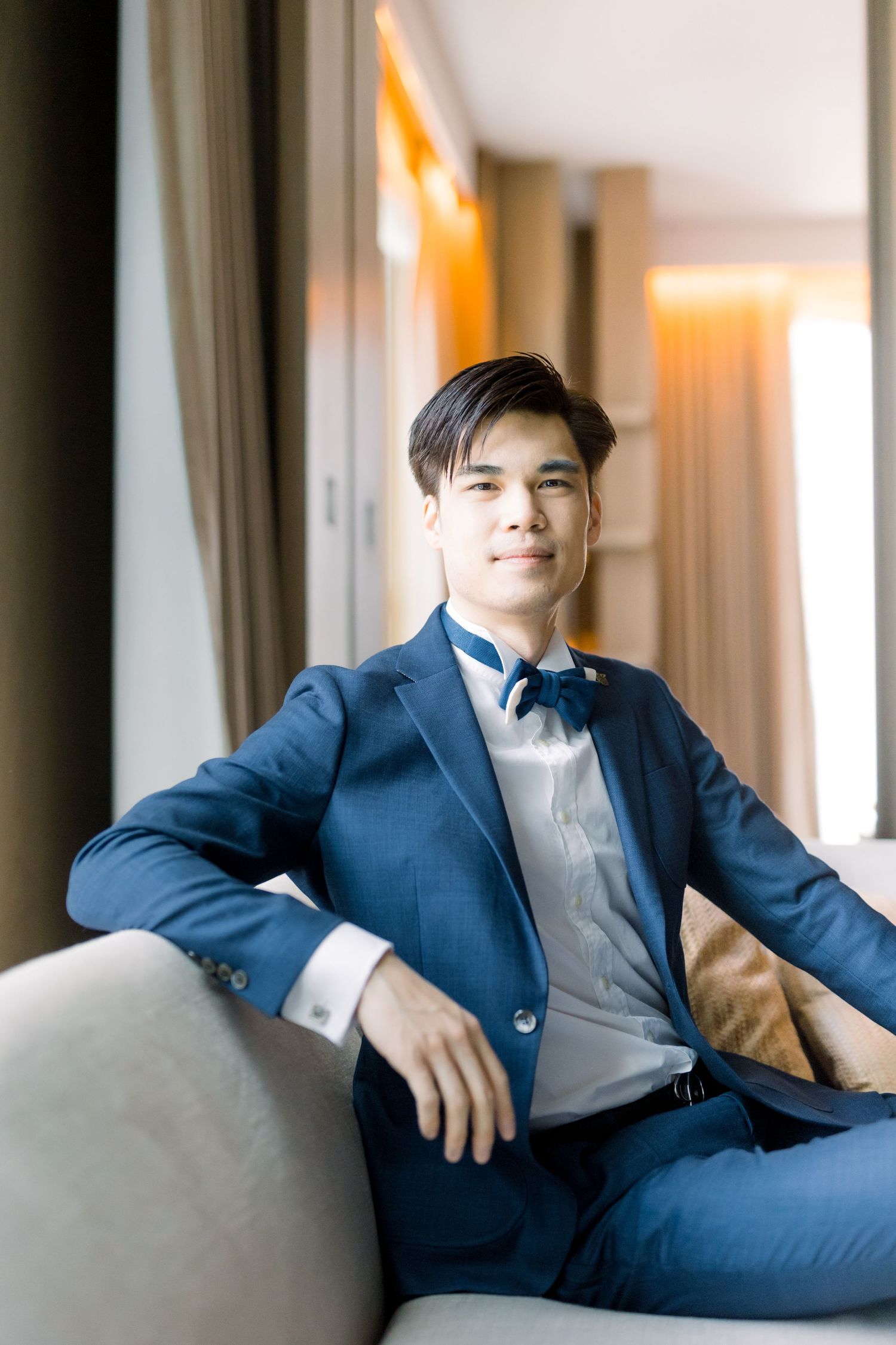 Groom lounges confidently on sofa in classic navy suit and bow tie.