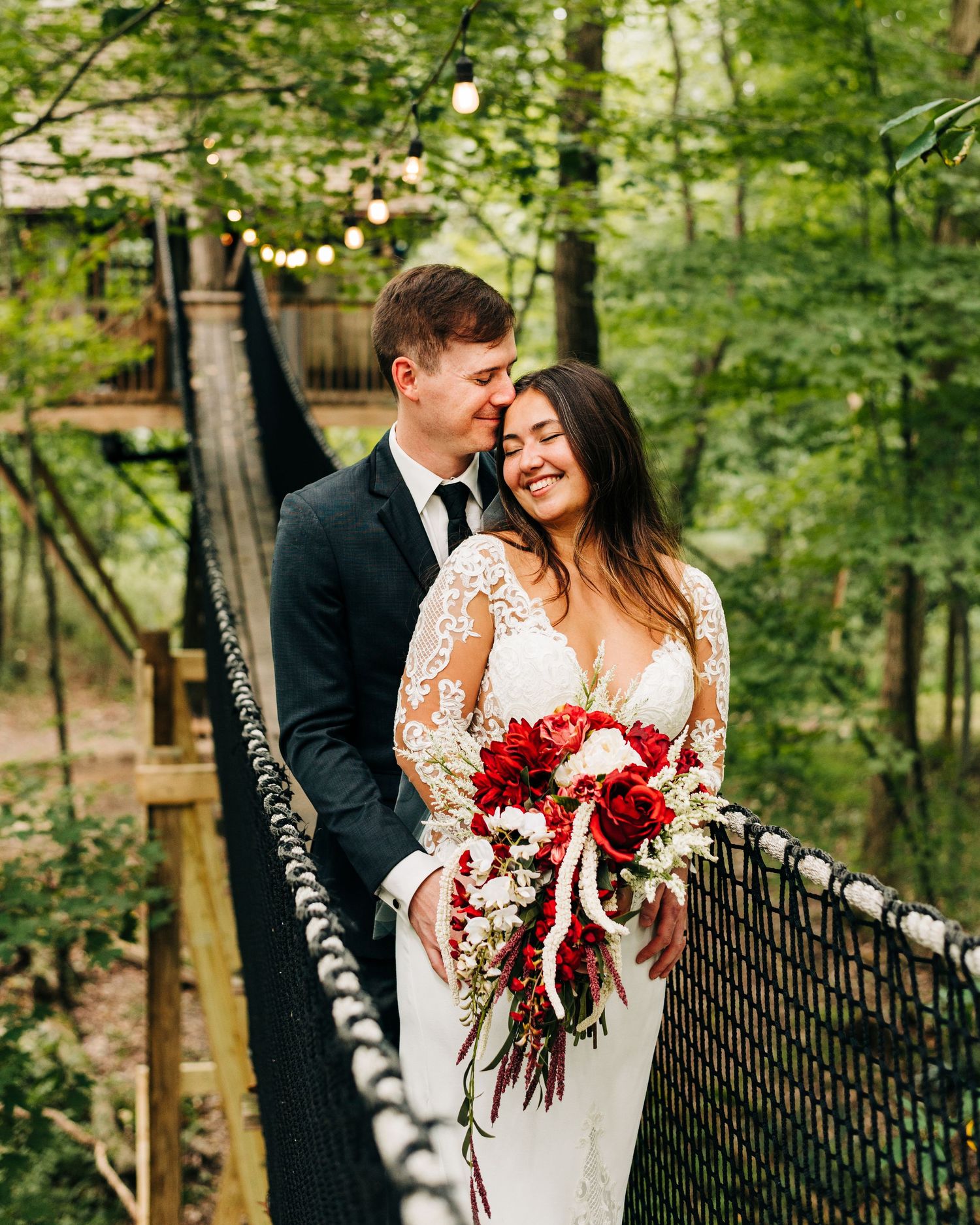 Couple sharing intimate moment on forest bridge with bride holding red bouquet.