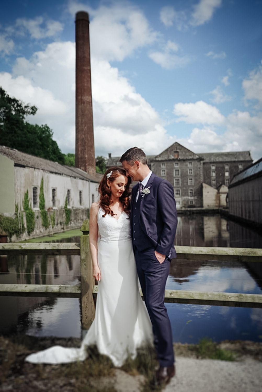 Couple posed by historic industrial building with brick chimney and reflective water feature.