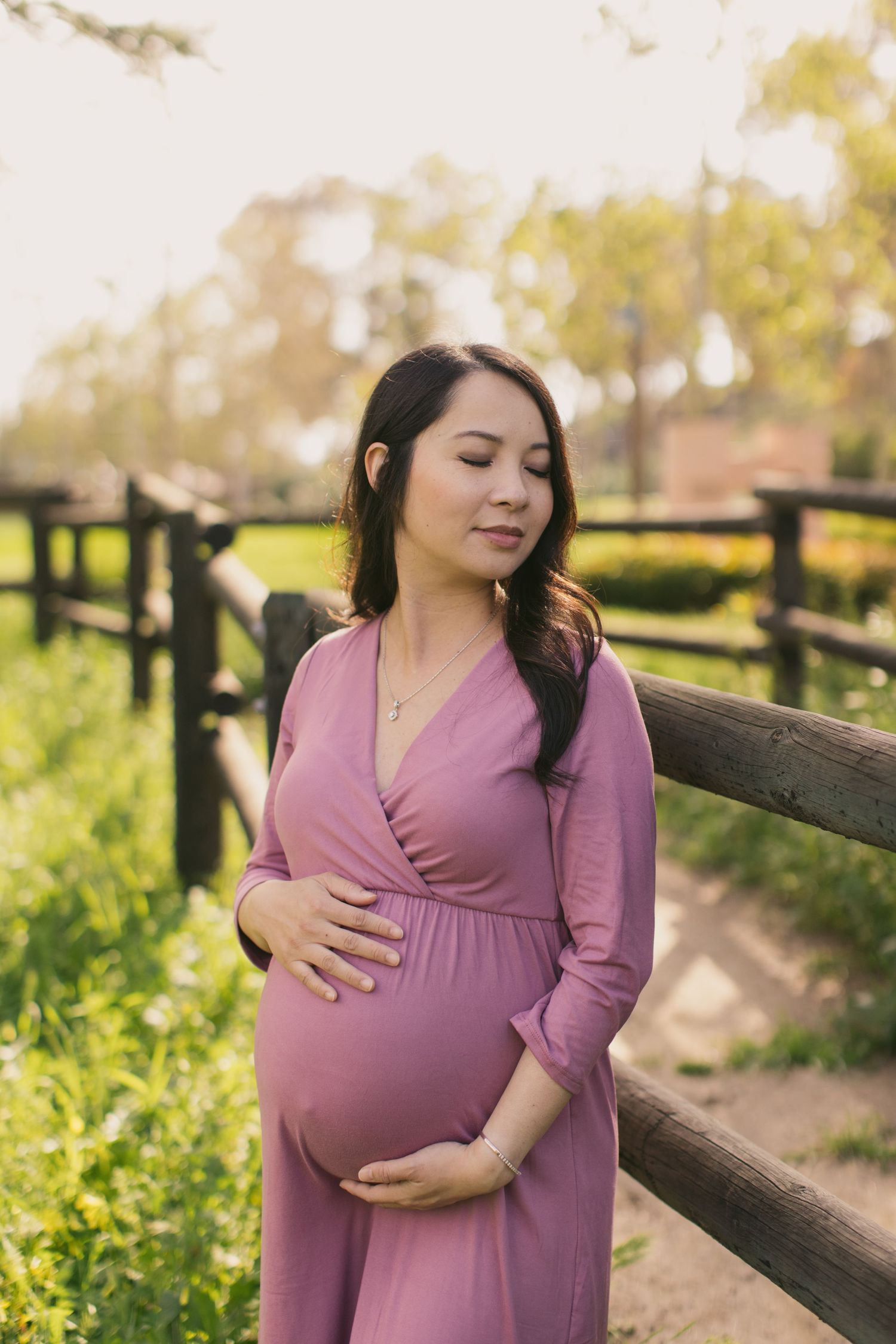 Maternity photo session in a park with a woman in a mauve dress standing by a wooden fence surrounded by yellow flowers.