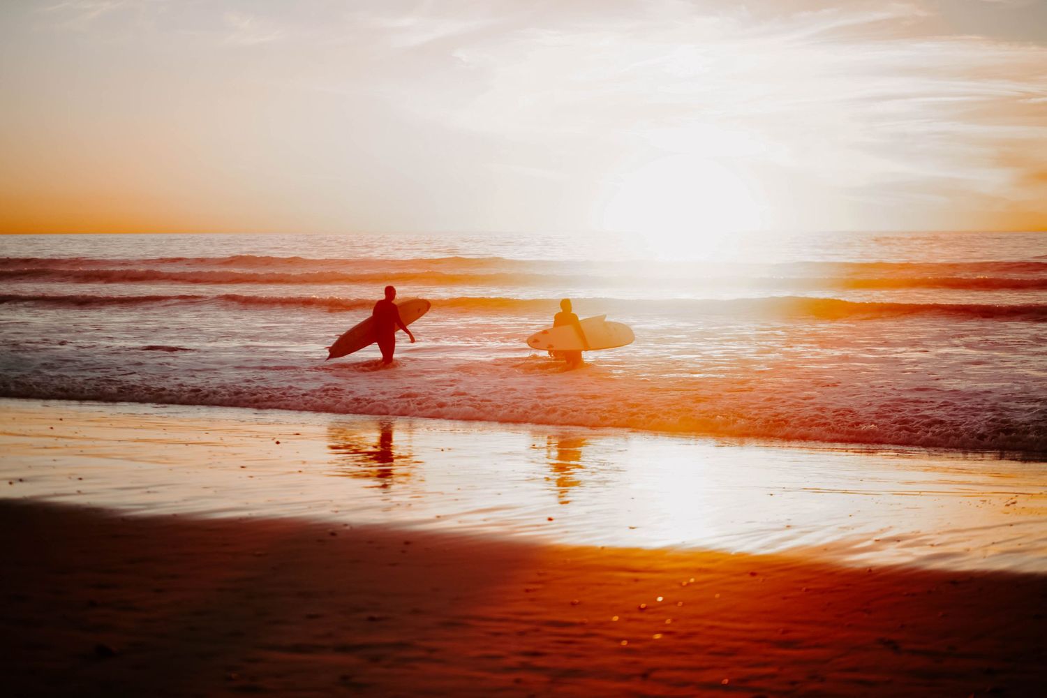 Golden hour photo captured of two surfers walking into the water during a sunset.