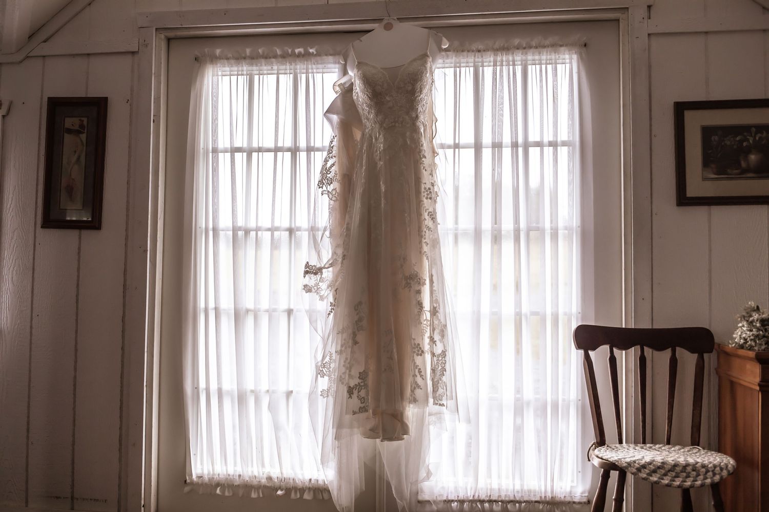Elegant white wedding dress hanging in front of sunlit windows in a vintage room.
