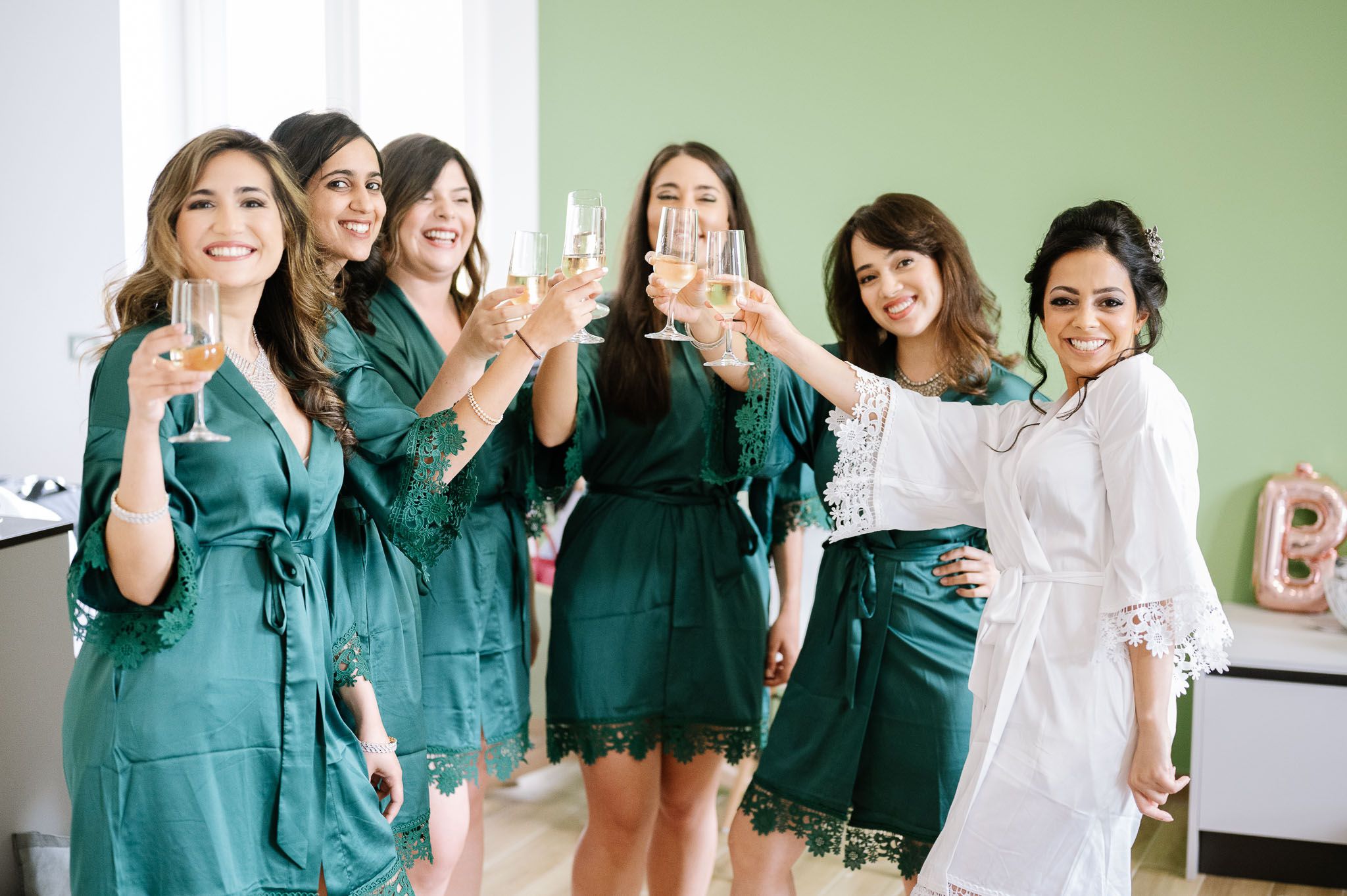 Group wearing emerald green robes toasting with champagne glasses during wedding preparations.
