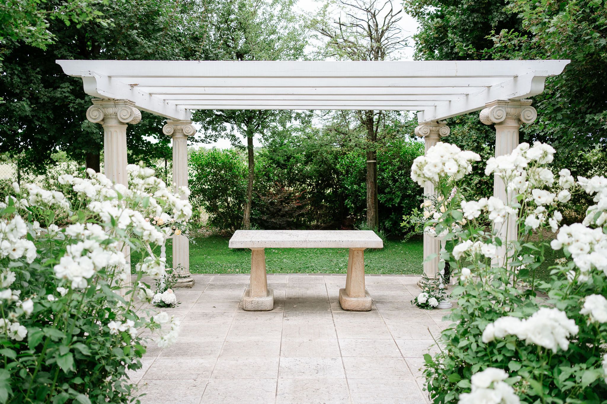 A white garden pergola with stone columns and climbing roses creates an elegant outdoor seating area.