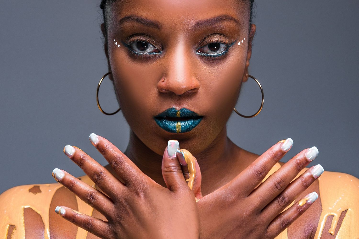 Beauty portrait showcasing vibrant teal lipstick, silver hoops, and artistic manicure against grey background.