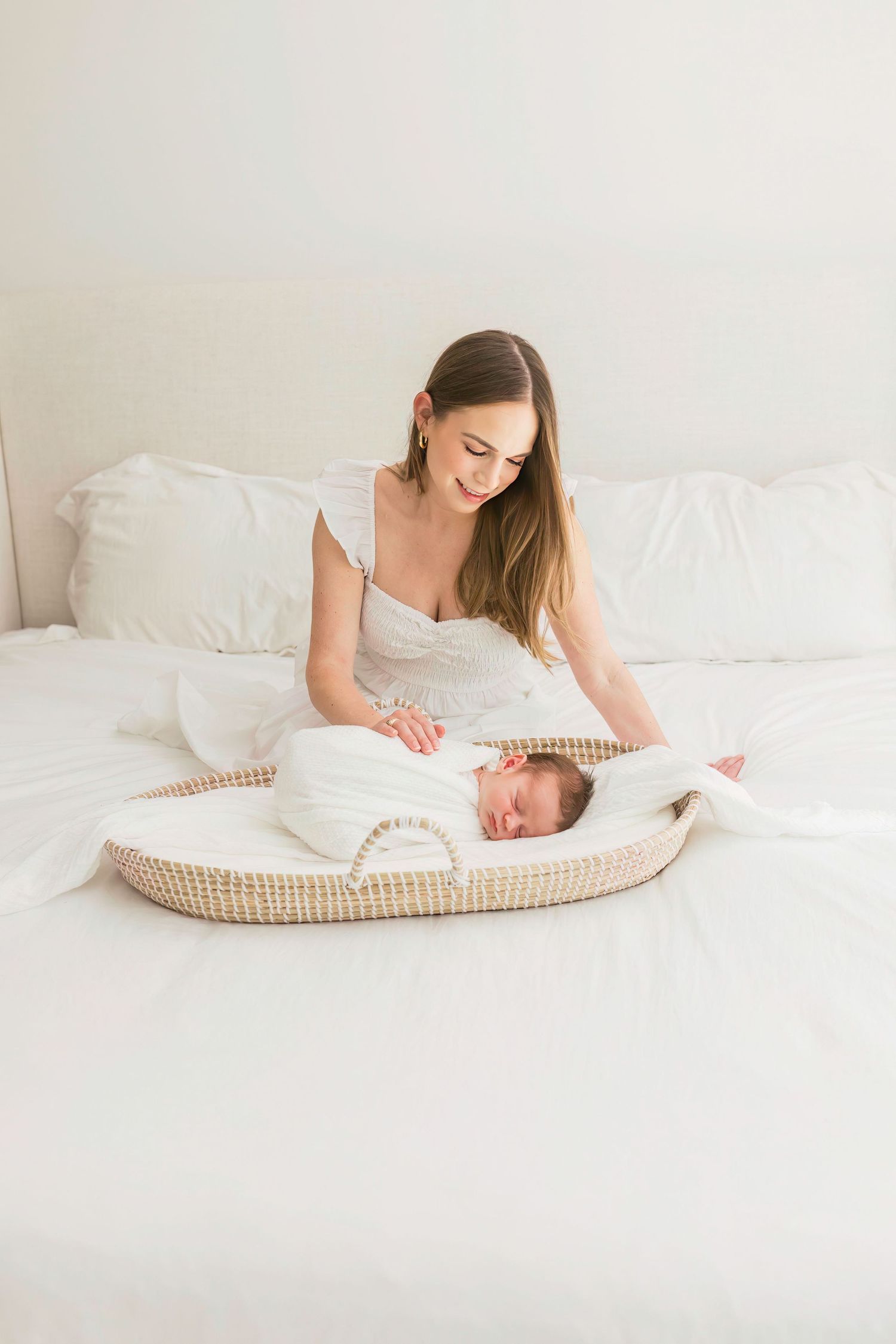 Mother sitting on white bed watching over baby in woven bassinet.