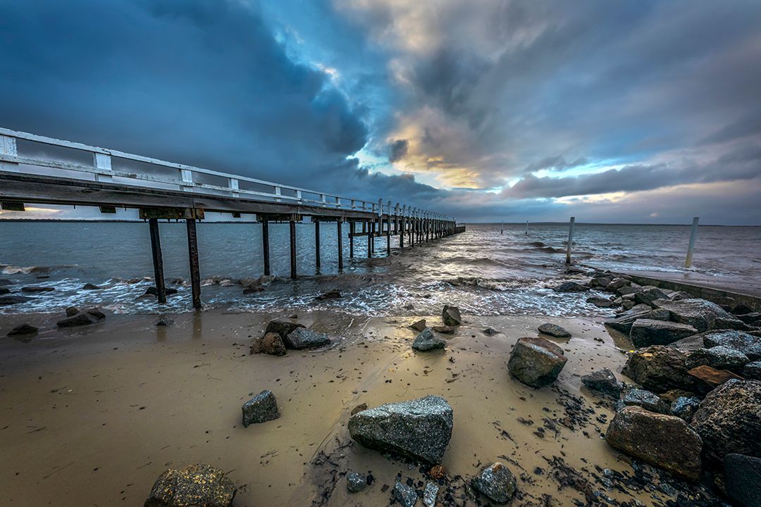 Dramatic cloudy sky over a wooden pier with rocky beach at low tide.