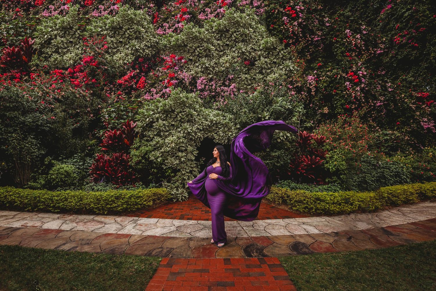 A dancer in a flowing purple dress poses gracefully in a garden with red flowers and greenery.