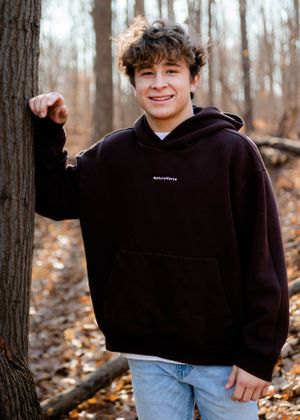 Person in black hoodie standing by tree trunk in fall forest setting.