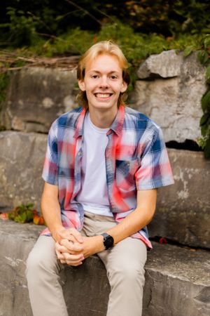 Student in plaid shirt and khakis sitting casually on stone wall in outdoor setting.