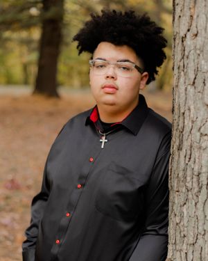 Person in black clergy shirt with cross necklace poses against tree in autumn setting.