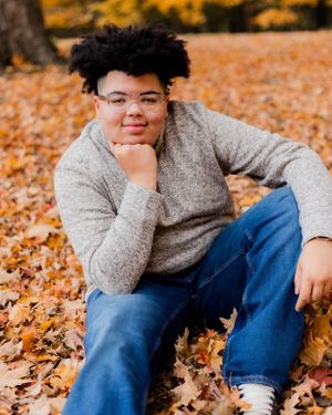 Individual in gray sweater and jeans sits among fallen autumn leaves.